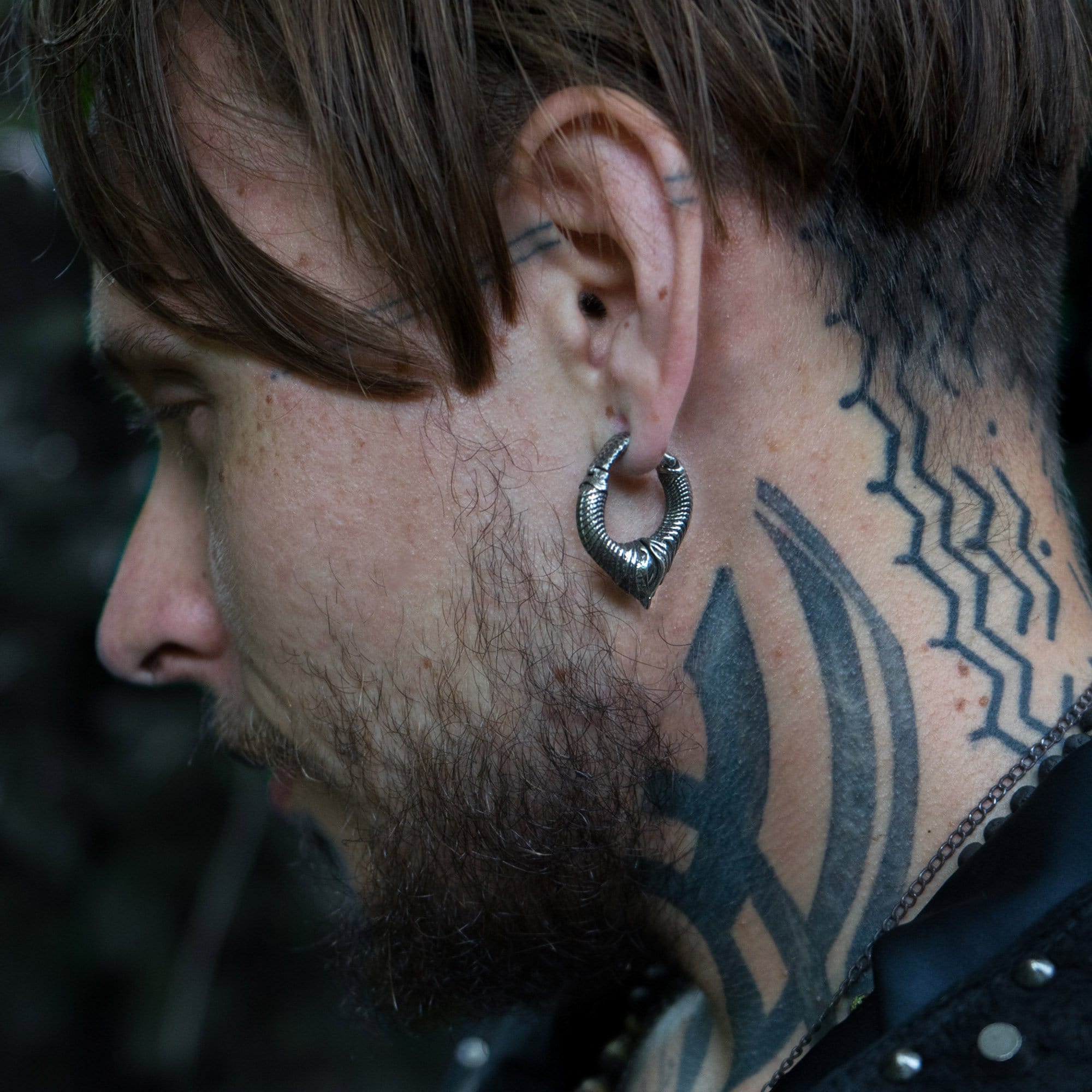A side profile of a person with brown hair and a beard wearing MARVDASHT Small Biomechanical Clicker Ear Weights in 925 Silver from Kaya Movement, designed for 2 gauge ears and shaped like serpents. Their neck and the side of their face feature multiple tattoos, including geometric patterns and shapes, enhancing the sci-fi vibes. The person is dressed in dark clothing.